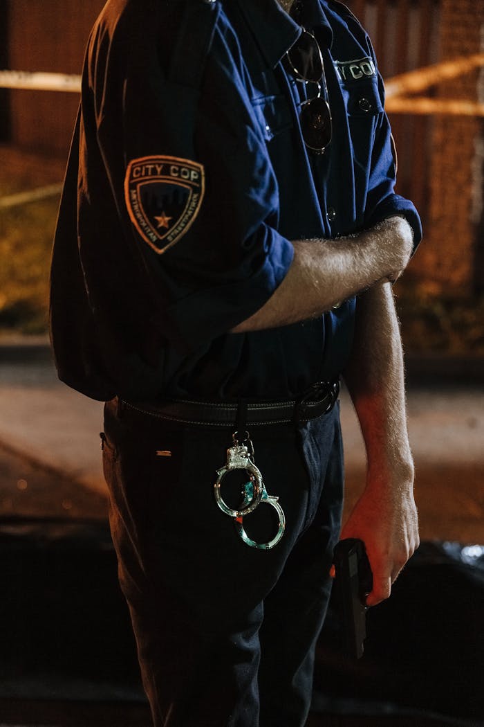 Police officer holding a gun with handcuffs hanging on belt in an outdoor setting.