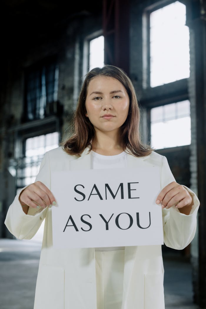 Woman in white coat holding 'Same As You' sign in industrial setting, promoting equality.