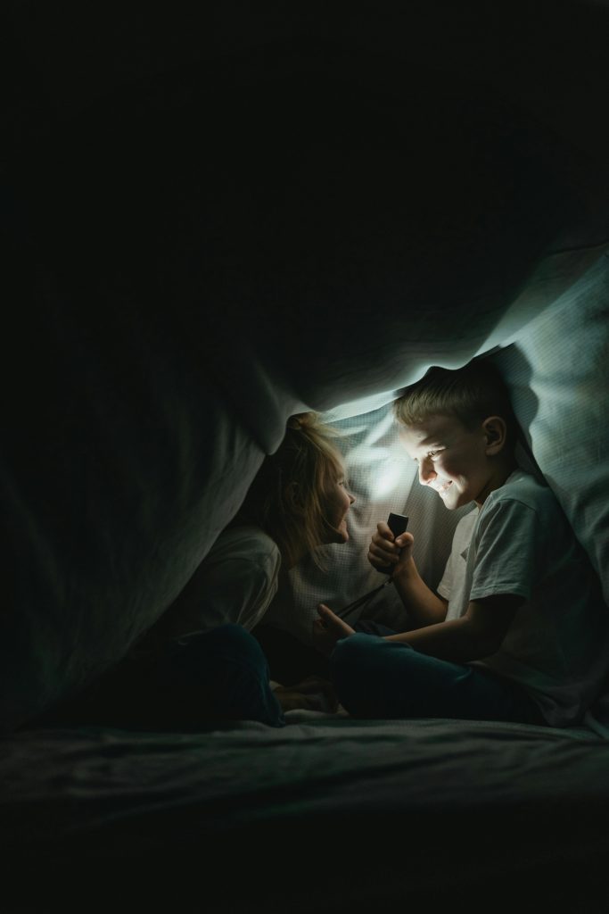 Two kids enjoying a playful moment with a flashlight inside a blanket fort.