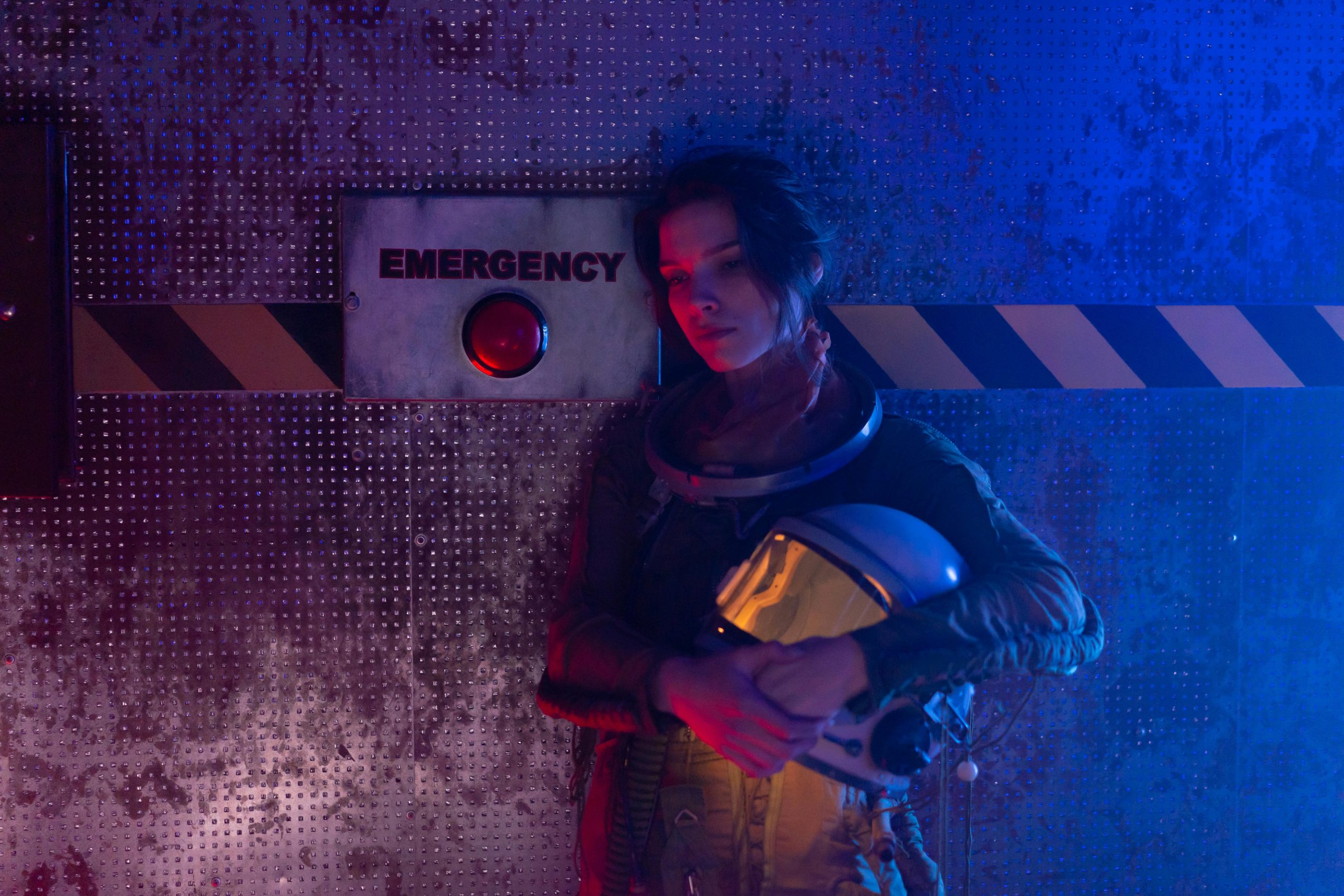 Young astronaut holding helmet in a dark emergency-themed room with red and blue lighting.