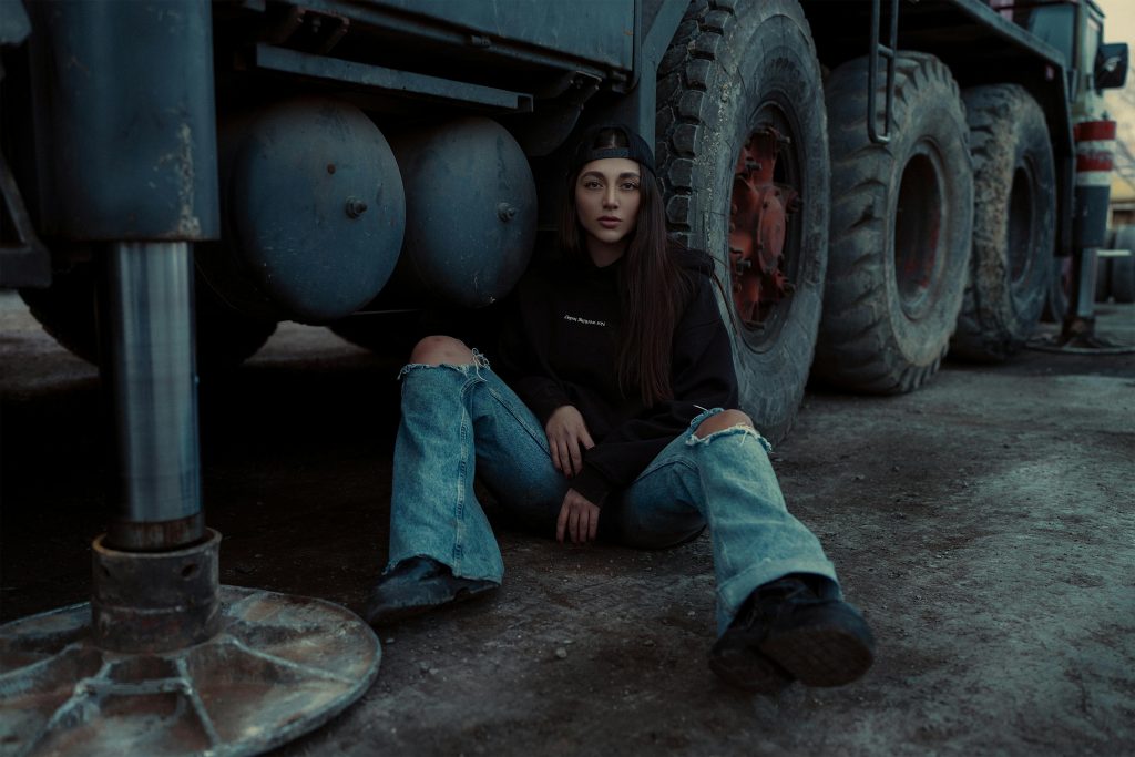 Young woman in casual fashion sitting by a large truck outdoors.