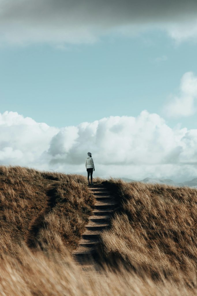 A lone woman hikes a grassy hill trail under a cloudy sky, epitomizing solitude.