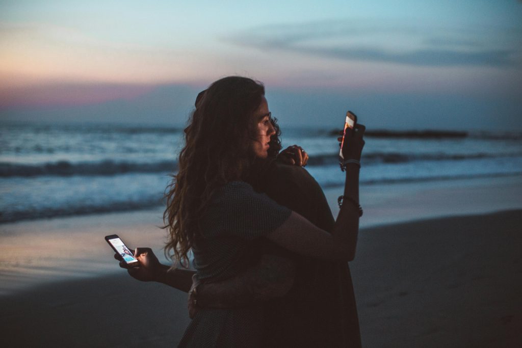 Couple embracing on a beach at sunset, using mobile phones, symbolizing modern love.