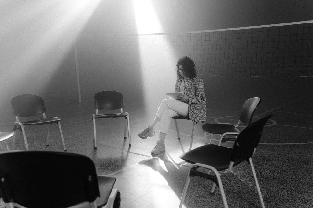 A solitary woman sits in a smoky, sunlit room surrounded by empty chairs, conveying a sense of solitude.