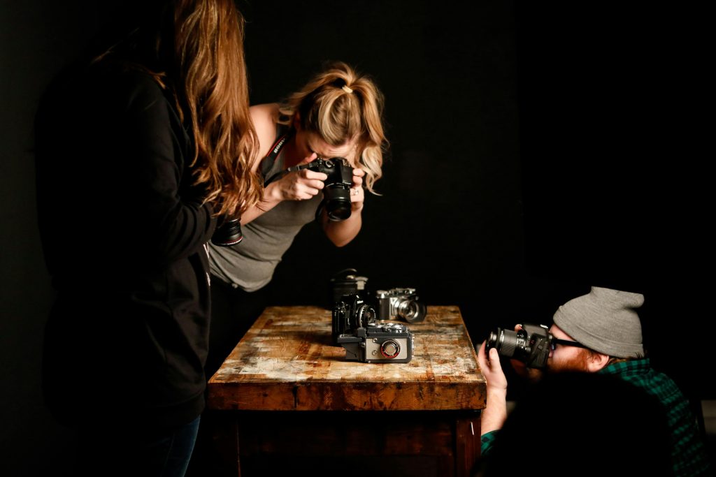 Three people use modern cameras to photograph vintage cameras on a wooden table indoors.