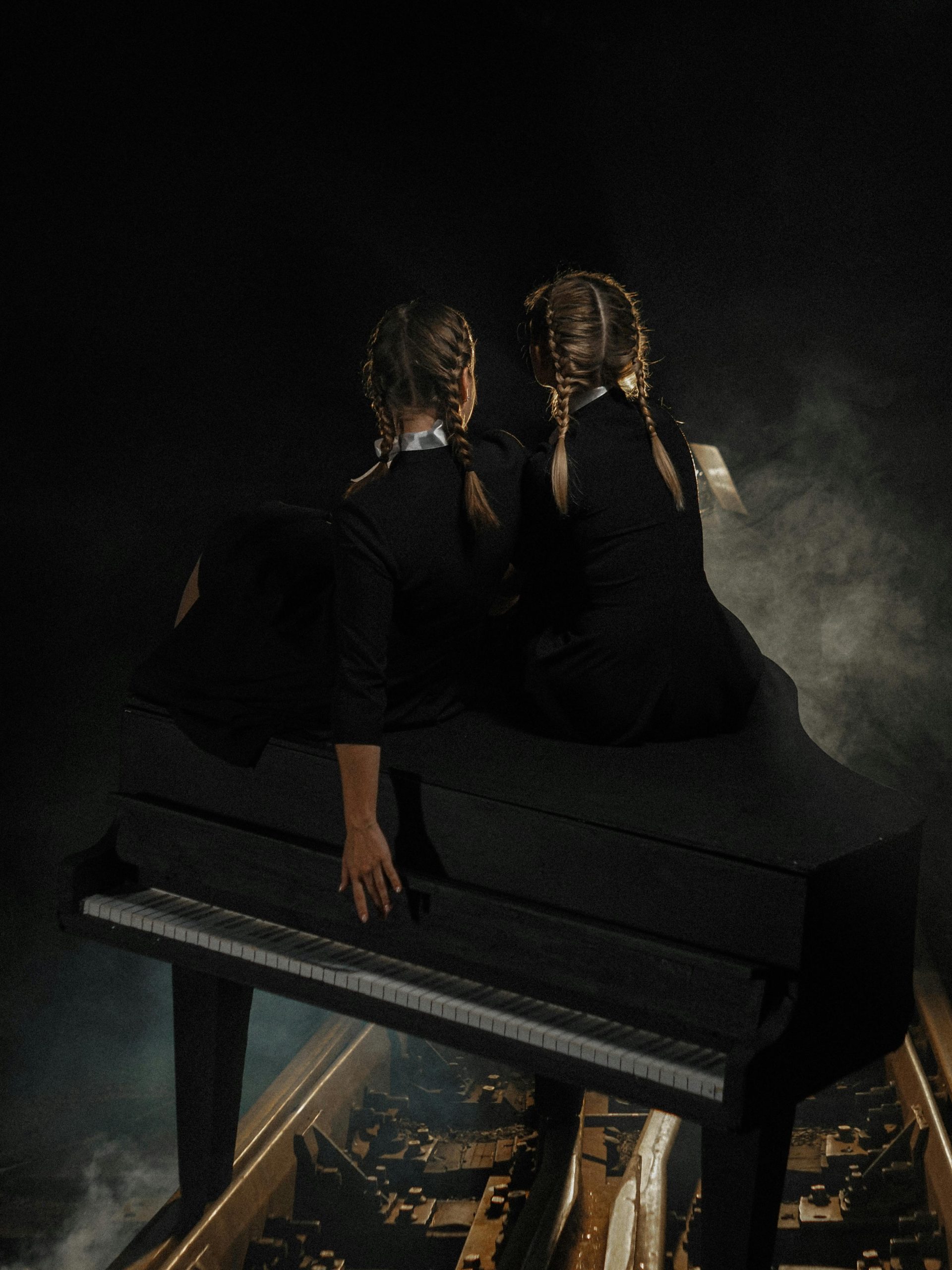 Two children in braids sit on a piano in a dark, smoky studio setting.