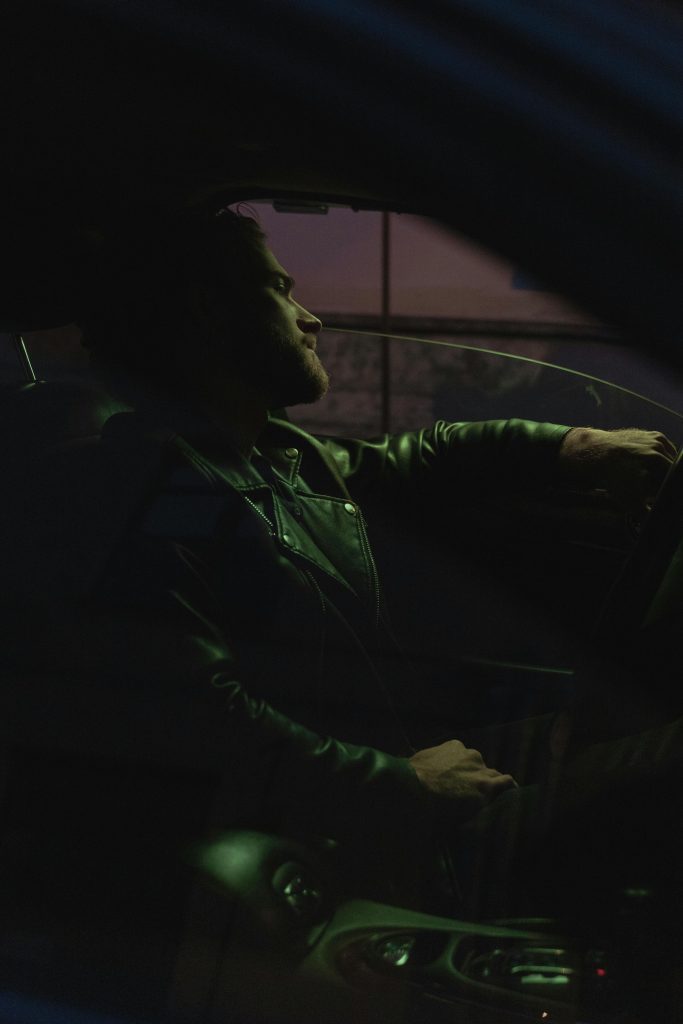 A man in a leather jacket sits in a car, driving at night, with a moody atmosphere.